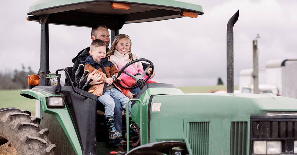 A father drives a green tractor with his two smiling children sitting beside him on a cloudy day, representing rural family life and the hardworking communities that chiropractic care supports.